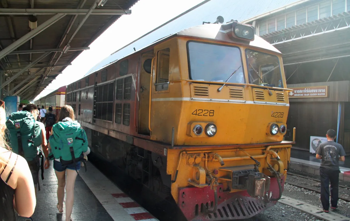 Backpackers walking along a station platform beside a yellow State Railway of Thailand diesel locomotive