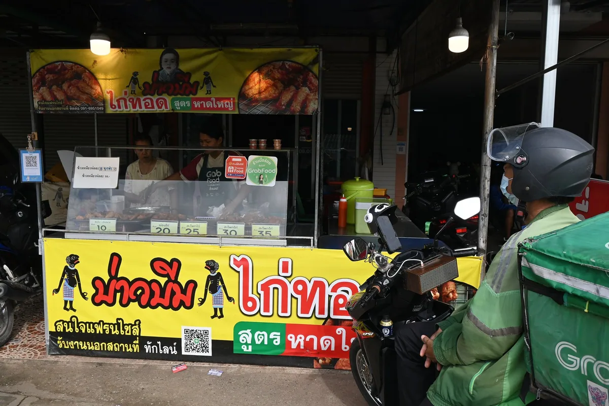 A Grab delivery rider in a green jacket waits on his motorbike beside a Thai fried-chicken stall