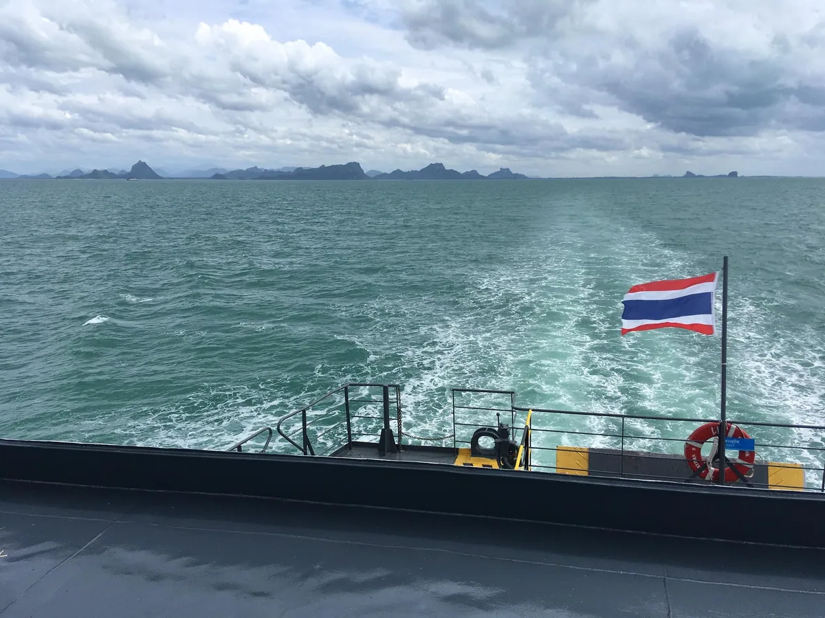 Thai flag flying over a ferry's wake in the Gulf, with the limestone hills of the Don Sak mainland coast on the horizon