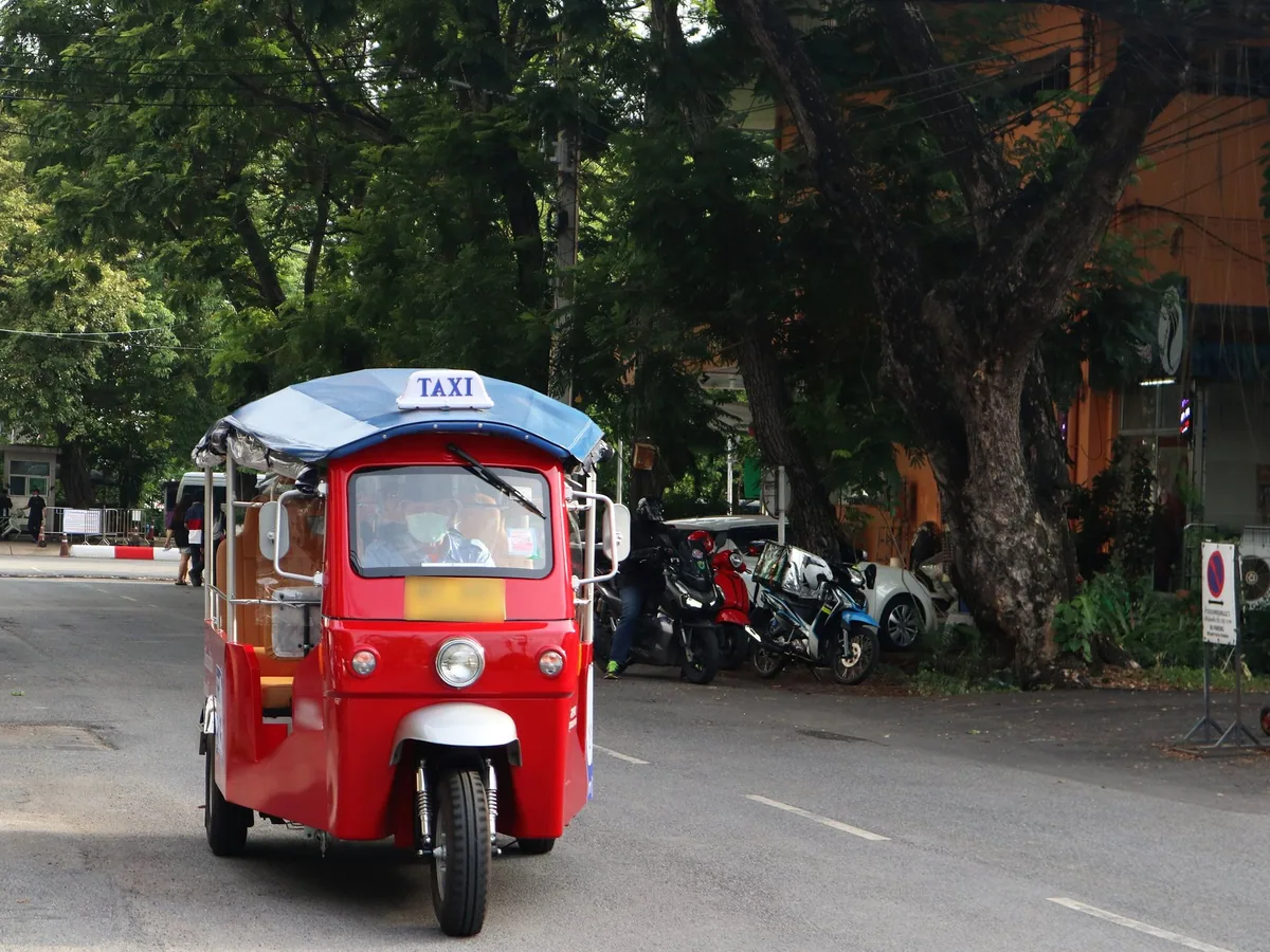 A red electric Muvmi tuk-tuk with a blue canopy and taxi sign driving down a tree-shaded Bangkok street