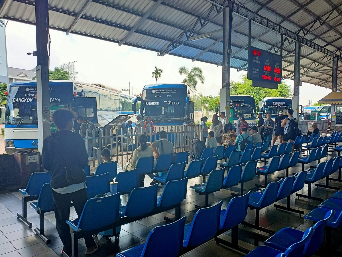 Travellers waiting on rows of blue seats under the roof of Pattaya's North Road bus station, with coaches parked behind