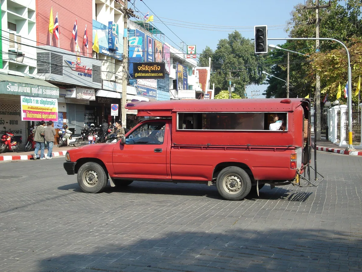 A red songthaew pickup with a covered passenger bench crossing a street in Chiang Mai