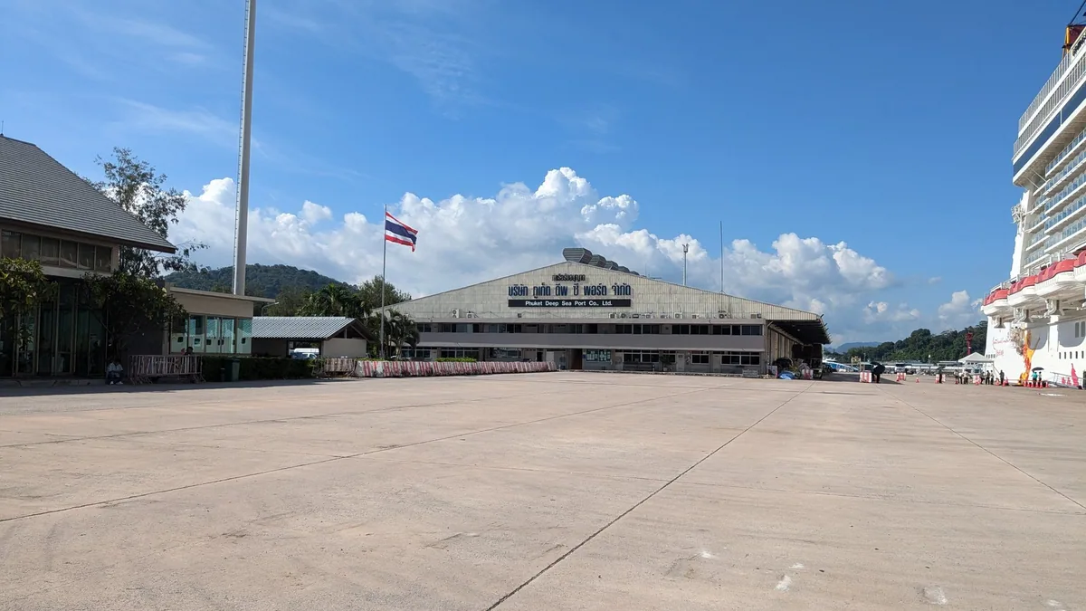 Thai flag over the terminal building of Phuket Deep Sea Port, with a berthed cruise ship's hull on the right