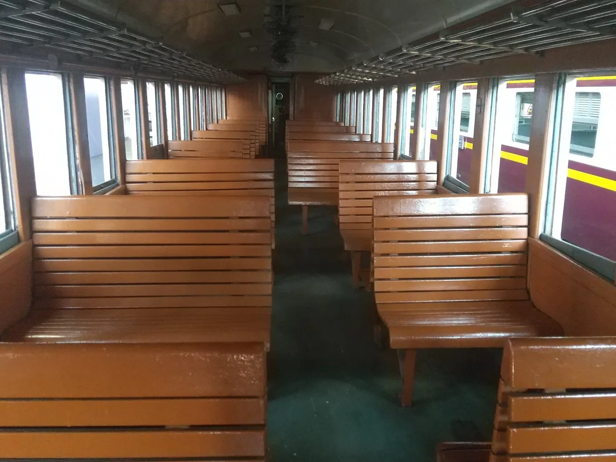 Rows of varnished wooden bench seats and open windows inside a Thai third-class railway carriage